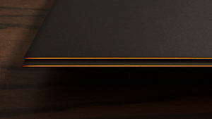 Extreme macro of a razor-thin matte black device edge on dark walnut catching a warm amber specular highlight, focusing on the side rail where a power button sits