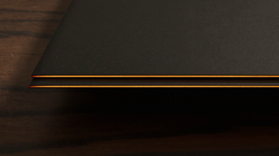 Extreme macro of a razor-thin matte black device edge on dark walnut catching a warm amber specular highlight, focusing on the side rail where a power button sits